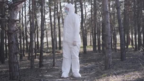 Joven caucásico consciente en traje blanco de seguridad de pie en el bosque junto a la carretera suburbana y mirando hacia otro lado sosteniendo la mano en la frente. El control de la cuarentena del coronavirus mide el cumplimiento. Amplio tiro . — Vídeos de Stock