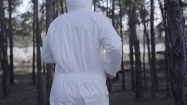 Vista posterior del joven hombre caucásico en traje de seguridad blanco, gafas protectoras y máscara facial corriendo en el soleado parque de primavera. Estilo de vida del deportista durante la cuarentena del coronavirus. Pandemia, Covid-19 . — Vídeos de Stock