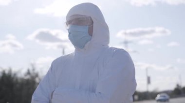 Açık havada duran güvenlik kıyafetli virolojistin yakın plan portresi. Kenar mahallede beyaz koruyucu giysiler, gözlükler ve yüz maskesi takan kendine güvenen genç bir adam. Covid-19, karantina, yaşam tarzı.