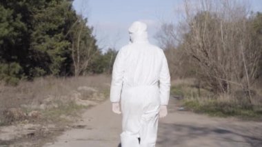 Beyaz takım elbiseli ve yüz maskeli bir erkek virüs uzmanı parktaki sokakta yürüyor. DSÖ 'nün Covid-19 karantinası hakkındaki tavsiyelerine uymayı kontrol eden bir adam dışarıda dolaşıyor..
