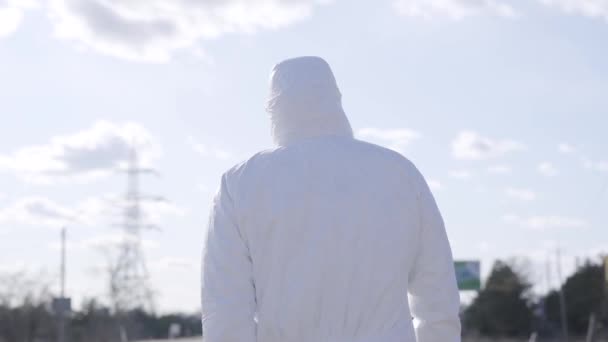 Vue arrière du jeune homme en costume de protection blanc marchant le long d'une route à moitié vide par une journée ensoleillée. Mode de vie urbain pendant la quarantaine de Covid-19. Europe centrale et orientale, politique de la santé, politique de la santé . — Video