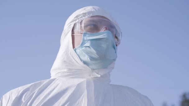 Detailní portrét mladého bělocha v bílém bezpečnostním obleku a ochranných brýlích na pozadí modré oblohy. Vážný virolog v masce na Covid-19 karanténě podle doporučení WHO. — Stock video