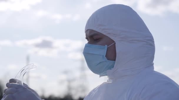 Detailní záběr mladého seriózního muže s ochrannými brýlemi. Sebevědomý běloch v bílém obleku a s obličejovou maskou tráví jarní den na předměstí. Koronavirová pandemie, Covid-19 karanténa. — Stock video