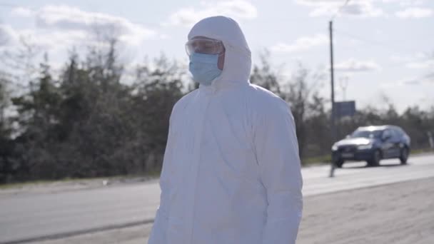 Sérieux jeune homme en combinaison de protection blanche debout sur la route de banlieue. Virologue caucasien avec masque facial et lunettes croisant les mains et détournant les yeux. Covid-19 mode de vie de quarantaine pandémique . — Video