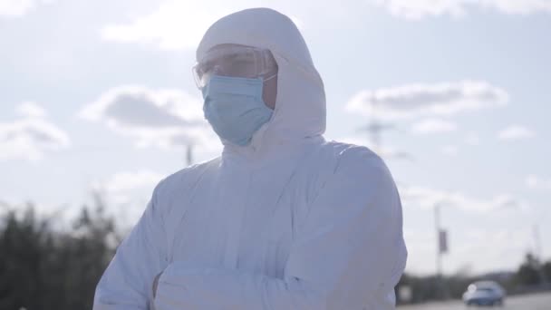 Close-up portrait of virologist in safety suit standing outdoors. Confident young man in white protective clothing, eyeglasses and face mask on suburban street. Covid-19, quarantine, lifestyle. — Stock Video