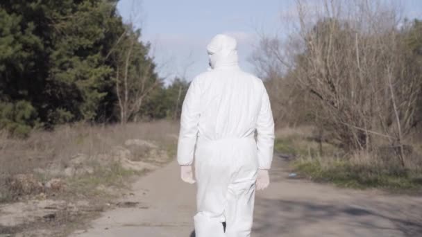 Vista posteriore del virologo maschile in tuta di sicurezza bianca e maschera facciale passeggiando lungo il vicolo nel parco. Uomo che passeggia all'aperto controllando il rispetto delle raccomandazioni dell'OMS sulla quarantena di Covid-19 . — Video Stock