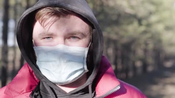 Gros plan portrait d'un jeune homme caucasien aux yeux gris portant un masque de protection à l'extérieur. Citoyen conscient prenant des mesures de sécurité se reposant dans le parc ensoleillé de printemps pendant la quarantaine Covid-19. Pandémie de coronavirus . — Video