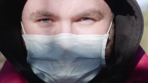 Extremo primer plano de la cara del joven hombre caucásico en la máscara en el día soleado al aire libre. Retrato del virólogo guapo confiado mirando la cámara. Covid-19, coronavirus, cuarentena, pandemia, estilo de vida . — Vídeo de stock