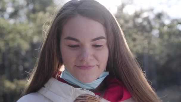 Närbild av brunett flicka tar av ansiktsmask och äter välsmakande croissant utomhus. Ung vit kvinna med mellanmål utomhus på Covid-19 karantän. Livsstil, coronaviruspandemi. — Stockvideo