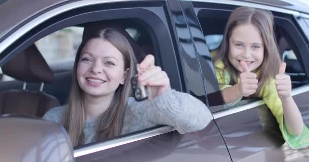 Mladá běloška se vychloubá klíčky od auta, zatímco malá holka ukazuje palce z auta. Bohatá obchodnice kupující nové vozidlo s dcerou v prodejně. Cinema 4k ProRes HQ. — Stock video