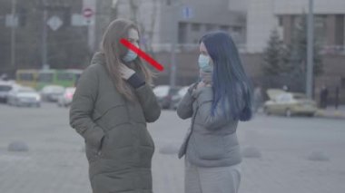 Koruyucu yüz maskesi takmış iki beyaz kadın dışarıda konuşuyorlar. Yasak haç ortaya çıkıyor ve 1.80 boyunda güvenlik işareti var. Arkadaştan 2 metre uzakta öksüren bir kız. Covid-19 karantina.
