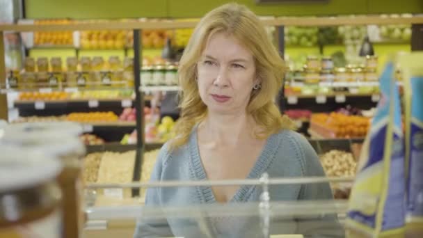 Portret van een volwassen blanke vrouw die door planken in de supermarkt kijkt. Geconcentreerde blonde huisvrouw die producten kiest in de supermarkt. Lifestyle, aankopen. levensmiddelen, kleinhandel. — Stockvideo
