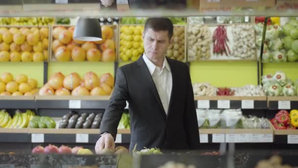 Серьезный кавказский мужчина в костюме выбирает фрукты киви в продуктовом магазине. Портрет уверенного взрослого бизнесмена, покупающего ягоды в розничном магазине. Потребительство, вегетарианство, здоровое питание, витаминная пища . — стоковое видео