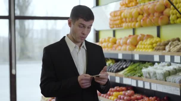 Porträt eines schockierten erwachsenen Mannes aus dem Kaukasus, der im Supermarkt durch die Kasse schaut. Brünettes Kerlchen im eleganten schwarzen Anzug überrascht mit Preisen im Einzelhandel. Konsum, Lebensstil, Einkaufen. — Stockvideo