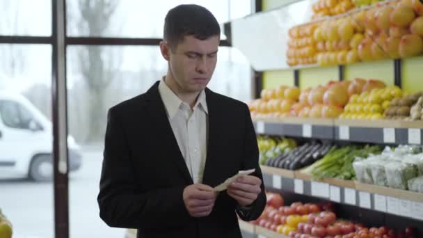 Porträtt av rika kaukasiska affärsman undersöka priset på mat i proposition och suckande. Ung man i kostym gör inköp i matvaror. Livsstil, konsumtion, shopping. — Stockvideo