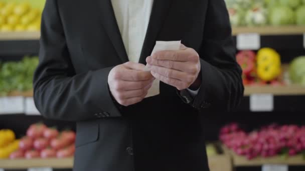 Vooraanzicht van de onherkenbare blanke man die de rekening in handen houdt en de prijs van het gekochte voedsel controleert in de supermarkt. Serieuze man onderzoekt de kosten van inkopen in de supermarkt. Levensstijl, financiën. — Stockvideo