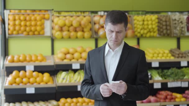 Vue de face de l'homme caucasien adulte vérifiant facture dans l'épicerie avec expression faciale surprise. Portrait d'un gars sérieux en costume regardant à travers les prix des aliments dans un supermarché. Finance, mode de vie . — Video