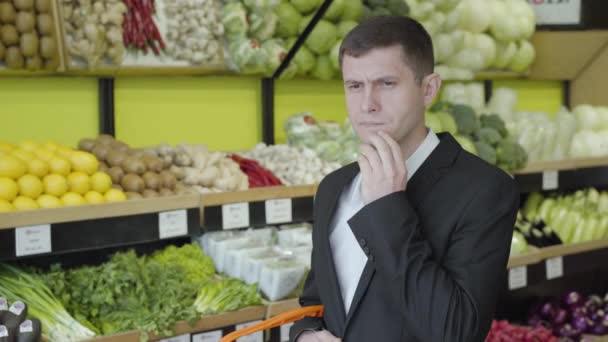 Retrato de morena caucasiano homem com olhos castanhos em pé no supermercado e pensamento. Cara séria em terno elegante decidir sobre variedade para comprar. Estilo de vida, consumismo, compras, varejo . — Vídeo de Stock