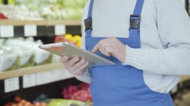 Raflarda yürüyüp tablet kullanan üniformalı tanınmamış bir erkek bakkal. Perakende dükkanındaki sebze ve meyve listesini kontrol eden yetişkin bir adam. Ticaret, tüketim, yaşam tarzı.