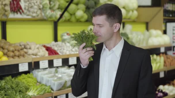 Porträt eines lächelnden kaukasischen Mannes, der frisches Grün im Lebensmittelladen riecht. Junger Mann im Anzug wählt Dill im Supermarkt. Kräuter, Gewürze, Kochen, Lebensstil. — Stockvideo