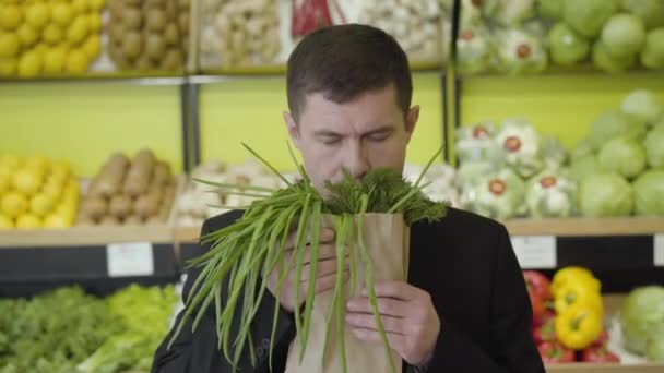 Framifrån av nöjda kaukasiska mannen luktar färska grönska gäng och sträcker den till kameran. Porträtt av leende man i elegant kostym köper hälsosam vegetarisk säsongsbetonad mat i mataffären. Livsstil — Stockvideo