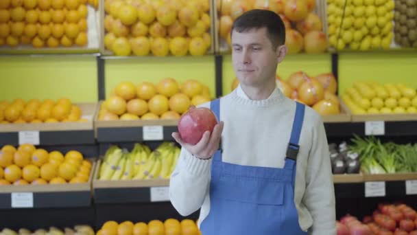 制服を着た正の白人ブルネットの男の肖像画はザクロを保持し、カメラで親指を表示します。食料品や笑顔に立つ自信のある労働者。商取引、健康的な食事、ビタミンフルーツ. — ストック動画