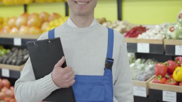Неузнаваемый улыбающийся белый мужчина, стоящий в продуктовом с папкой. Взрослый работник супермаркета в синей форме позирует в зеленом магазине. Торговля, профессия, образ жизни, бизнес . — стоковое видео