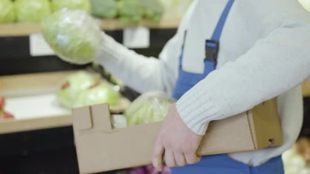 Employé masculin méconnaissable mettant des choux sur l'étagère dans l'épicerie et partant. Homme en uniforme ajoutant assortiment de légumes au supermarché. Commerce, commerce de détail, commerce, services . — Video