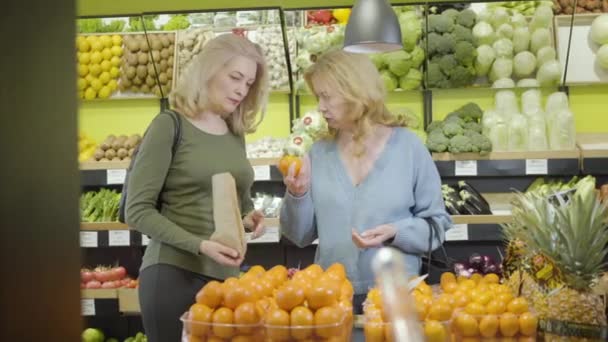 Två vuxna vita hemmafruar som plockar mandariner i mataffären. Seriösa äldre kvinnor som lägger frukt i papperspåse och pratar. Livsstil, vitamindiet, vegetarism, konsumism. — Stockvideo