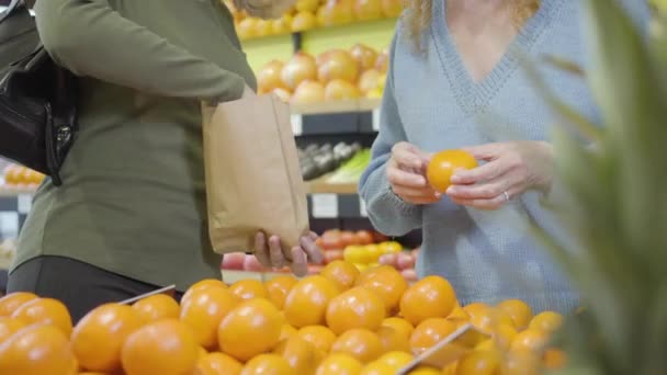 Två oigenkännliga kvinnliga kunder väljer tangeriner i mataffären. Vuxna kaukasiska kvinnor väljer färska ekologiska frukter i detaljhandeln. Hälsosam kost, vitaminmat, vegetarism, livsstil. — Stockvideo