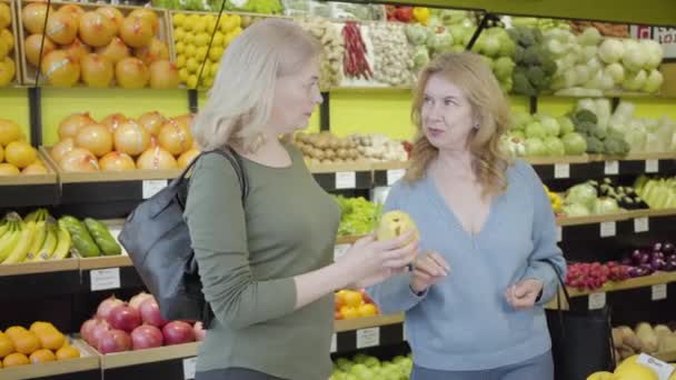 Deux femmes au foyer caucasiennes matures sentant la poire jaune fraîche à l'épicerie et parlant. Des femmes confiantes discutant de fruits frais juteux au supermarché. Achats, commerce, mode de vie, consumérisme . — Video