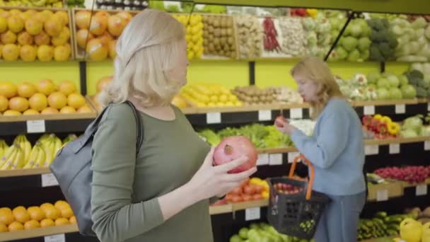 Portret van een volwassen blanke vrouw die met een vriend adviseert over de aankoop van sappige biologische granaatappel. Serieuze huisvrouwen die groenten en fruit kopen in de supermarkt. Gezond eten, levensstijl, handel. — Stockvideo