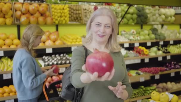 カメラにザクロを伸ばし、背景に果物の香りを嗅ぐ他の女性客として話す陽気なシニア白人女性。食料品で健康食品を購入する喜びのクライアントの肖像画. — ストック動画