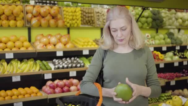 Porträtt av lycklig säker kaukasisk kvinna som köper frukt i mataffären. Senior blond hemmafru lägger pomelo i korgen och ler. Njutning, livsstil, shopping, konsumism. — Stockvideo