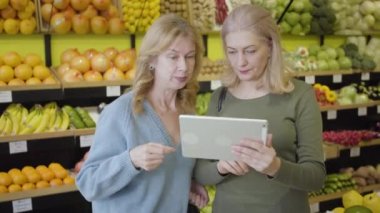 Markette tablet kullanan Kafkasyalı yaşlı kadınların portresi. İki ev hanımı mal ile konuşma arasında duruyor. Kablosuz teknoloji, yaşam tarzı, tüketim, alışveriş.