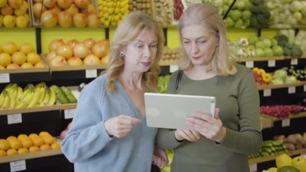 Portré pozitív kaukázusi idős nőkről, akik tablettát használnak a boltban. Két háziasszony áll sorok között és beszél. Vezeték nélküli technológia, életmód, fogyasztás, vásárlás. — Stock videók