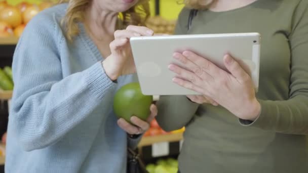 Две неузнаваемые кавказки используют планшет для проверки происхождения Помело в продуктовом магазине. Серьезные дамы делают покупки в супермаркете. Здоровое питание, образ жизни, консьюмеризм, диета . — стоковое видео