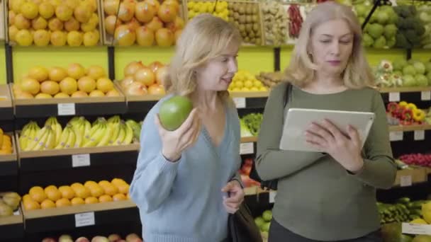 Tabletli kendine güvenen sarışın beyaz kadın, marketteki en iyi meyveyi seçmesini tavsiye ediyor. Olgun kız arkadaşlar marketten organik gıdalar alıyorlar.. — Stok video