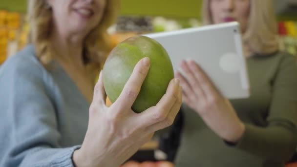 Gros plan de la main blanche femelle tenant pomelo comme des femmes floues en utilisant la tablette à l'arrière-plan. Aînées positives confiantes vérifiant l'origine des fruits à l'aide de technologies modernes . — Video