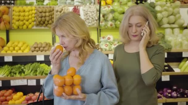 Schöne ältere kaukasische Frau, die frisch leckere Mandarinen riecht, während ihre Freundin am Telefon spricht. Positive Hausfrauen, die Obst im Lebensmittelladen kaufen. Lebensstil, Konsum, gesunde Ernährung. — Stockvideo
