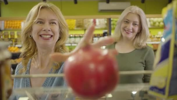 O foco muda de romã orgânica fresca suculenta deitada na prateleira no supermercado para rostos felizes de duas mulheres caucasianas seniores colocando-o no cesto de compras. Estilo de vida, consumismo, alimentação saudável . — Vídeo de Stock