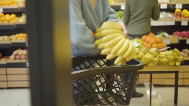 Eine bis zur Unkenntlichkeit erwachsene kaukasische Frau mit Einkaufskorb läuft durch die Reihen im Supermarkt und nimmt Früchte mit. Kundin kauft Bio-Bananen und Mandarinen. Lebensstil, vegane Ernährung, gesunde Ernährung. — Stockvideo