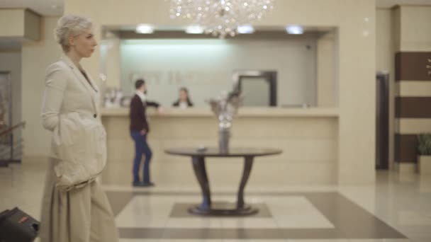 Zelfverzekerde blanke blonde vrouw met koffer passeren hotel vestibule met wazig mensen praten aan de receptie. Rijke elegante dame die accommodatie betreedt. Lifestyle, luxe, toerisme. — Stockvideo