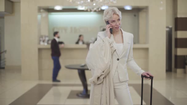 Молода стильна бізнес-леді в елегантному костюмі говорить по телефону в розкішному вестибюлі. Портрет щасливої впевненої жінки, що висить і посміхається, як стоячи в розкішному готелі. Туризм, спосіб життя . — стокове відео