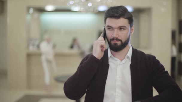 Guapo caucásico morena hombre con barba colgando teléfono y sonriendo a la cámara en el fondo de recepción borrosa en el lujoso hotel. Cliente varón confiado posando en el interior. Concepto turístico . — Vídeo de stock