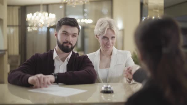 Portrait d'un homme caucasien positif signant des documents en tant que son conjoint prenant carte clé de chambre de la réceptionniste. Heureux couple marié s'enregistrant à l'hôtel de luxe. Des gens qui quittent le lobby de haut niveau . — Video