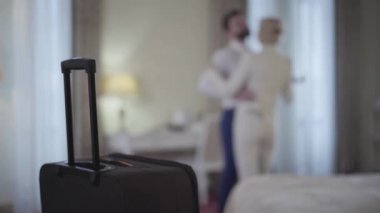 Arka planda dans eden, önde duran seyahat çantalı, bulanık, beyaz bir çift. Otel odasında vals yapan mutlu aşıklar. Turizm, aşk, yaşam tarzı, mutluluk.