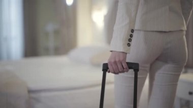 Otel odasında yatağa yaklaşan bavullu, tanımlanamayan beyaz kadının arka görüntüsü. Beyaz takım elbiseli zarif genç bayan tatil köyüne geliyor. Turizm, yaşam tarzı, seyahat, iş gezisi.