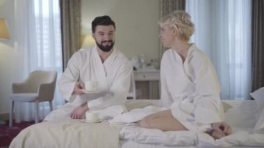 Bornozlu mutlu erkek ve kadın otel odasında çay fincanı tokuşturuyor. Genç Kafkasyalı karı ve koca üst sınıf konaklamalarda dinleniyor. Turizm, tatiller, yaşam tarzı, boş zaman.