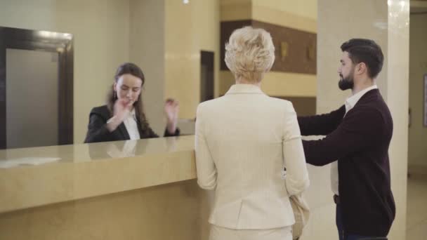 Young Caucasian female receptionist giving documents for sign to man and room room key card to woman. Wealthy couple of clients leaving reception desk. Arrival, tourism, service, luxury, lifestyle. — Stock Video